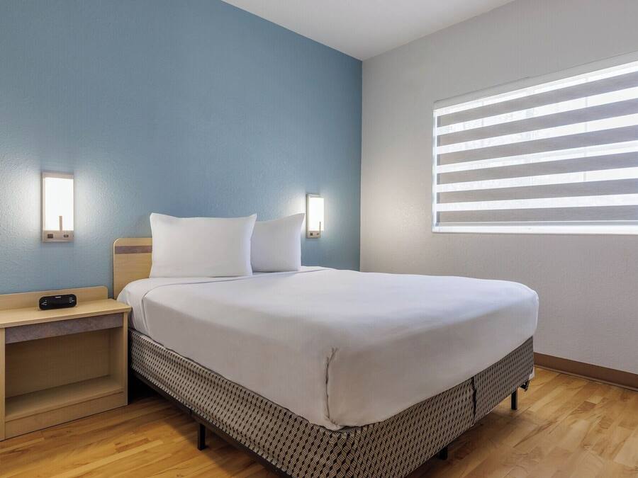 Ein Hotelzimmer mit Bett, Nachttischen, einem Fenster mit Jalousien und einer Wandleuchte.