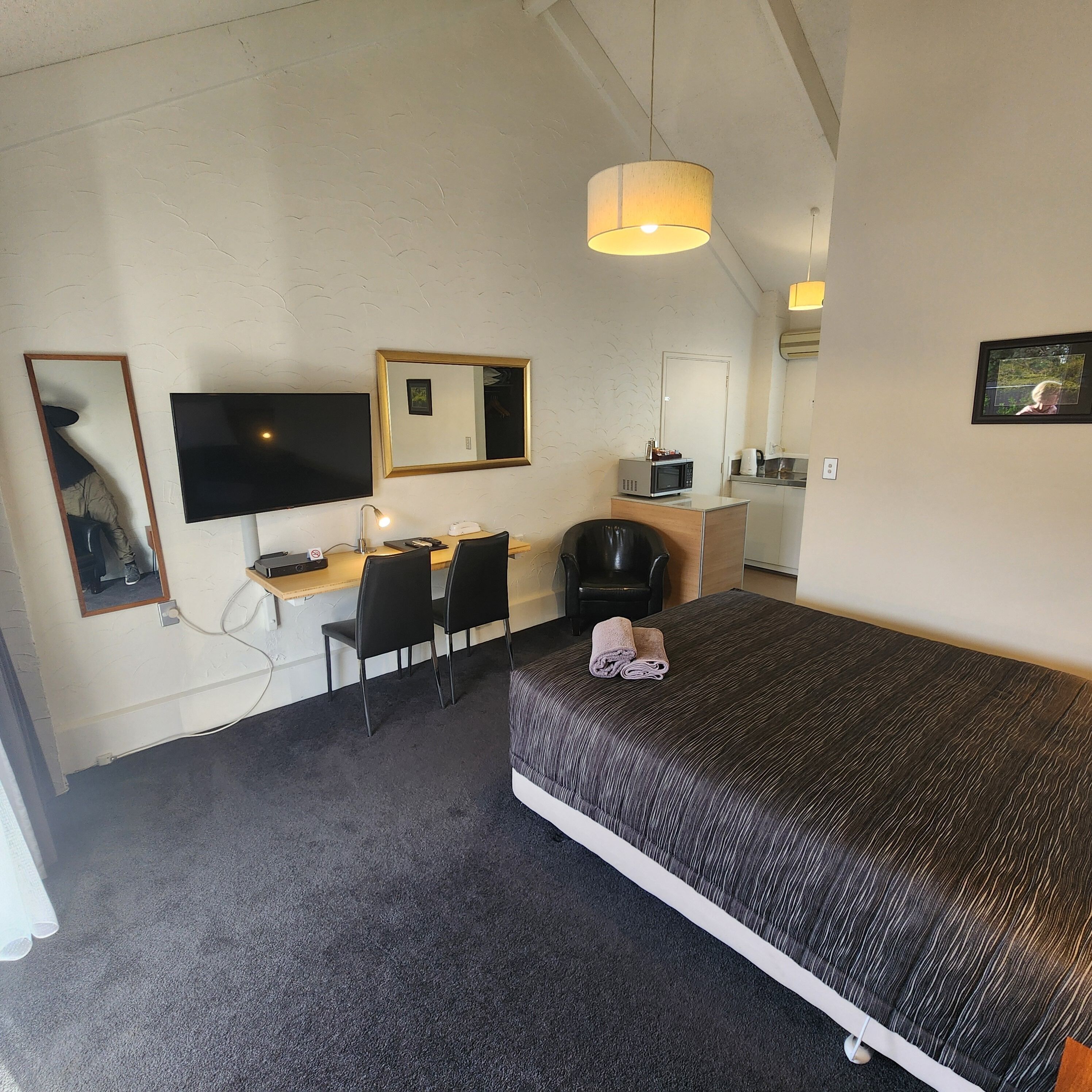 Habitación de hotel compacta con cama, escritorio con silla, televisor, microondas y una pequeña kitchenette.