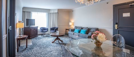 A hotel room with a glass dining table, a sofa with blue cushions, a TV, and a chandelier.