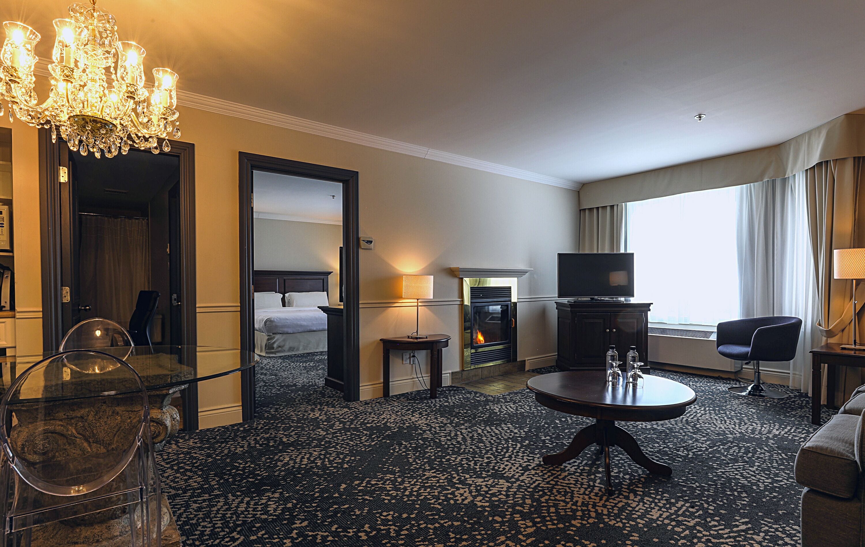 Hotelkamer met een kroonluchter, een open haard, een televisie en een groot raam met gordijnen.