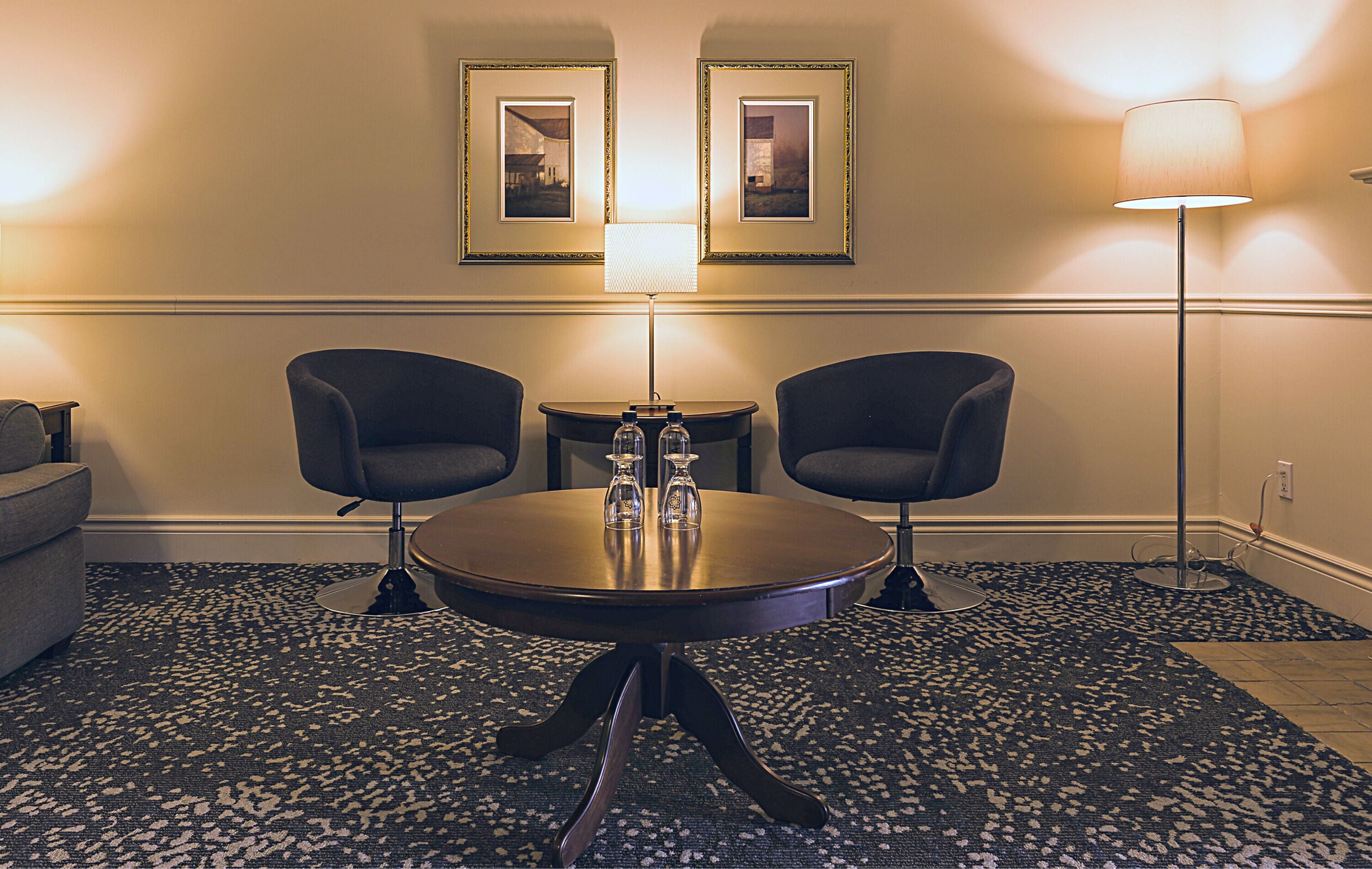 Ein Hotelzimmer mit rundem Tisch, zwei Sesseln, einer Stehlampe und zwei gerahmten Bildern an der Wand.