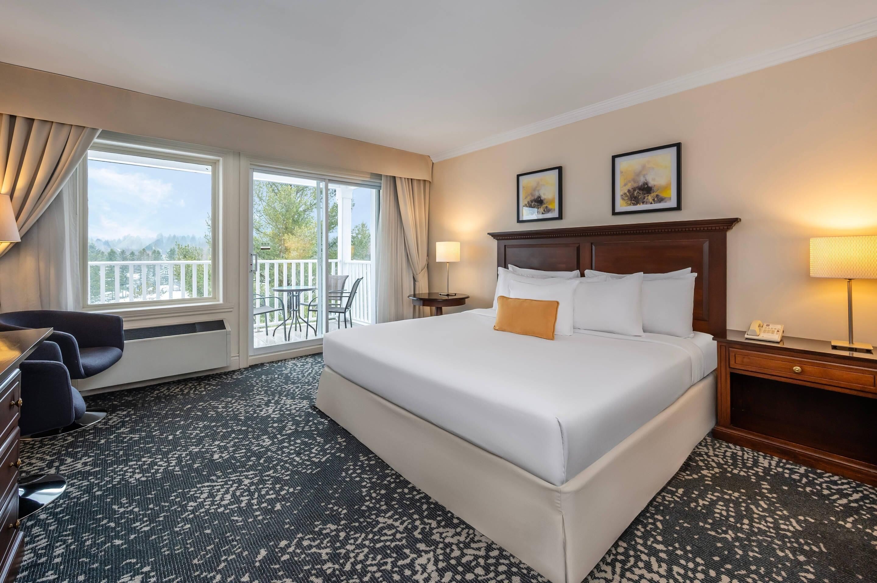 A hotel room with a large bed, a desk, a chair, a balcony with a view, and two framed pictures on the wall.
