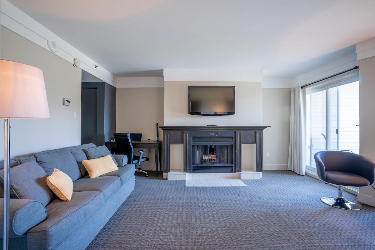 Ein modernes Wohnzimmer mit grauem Sofa, Flachbildfernseher, Kamin und großem Fenster.