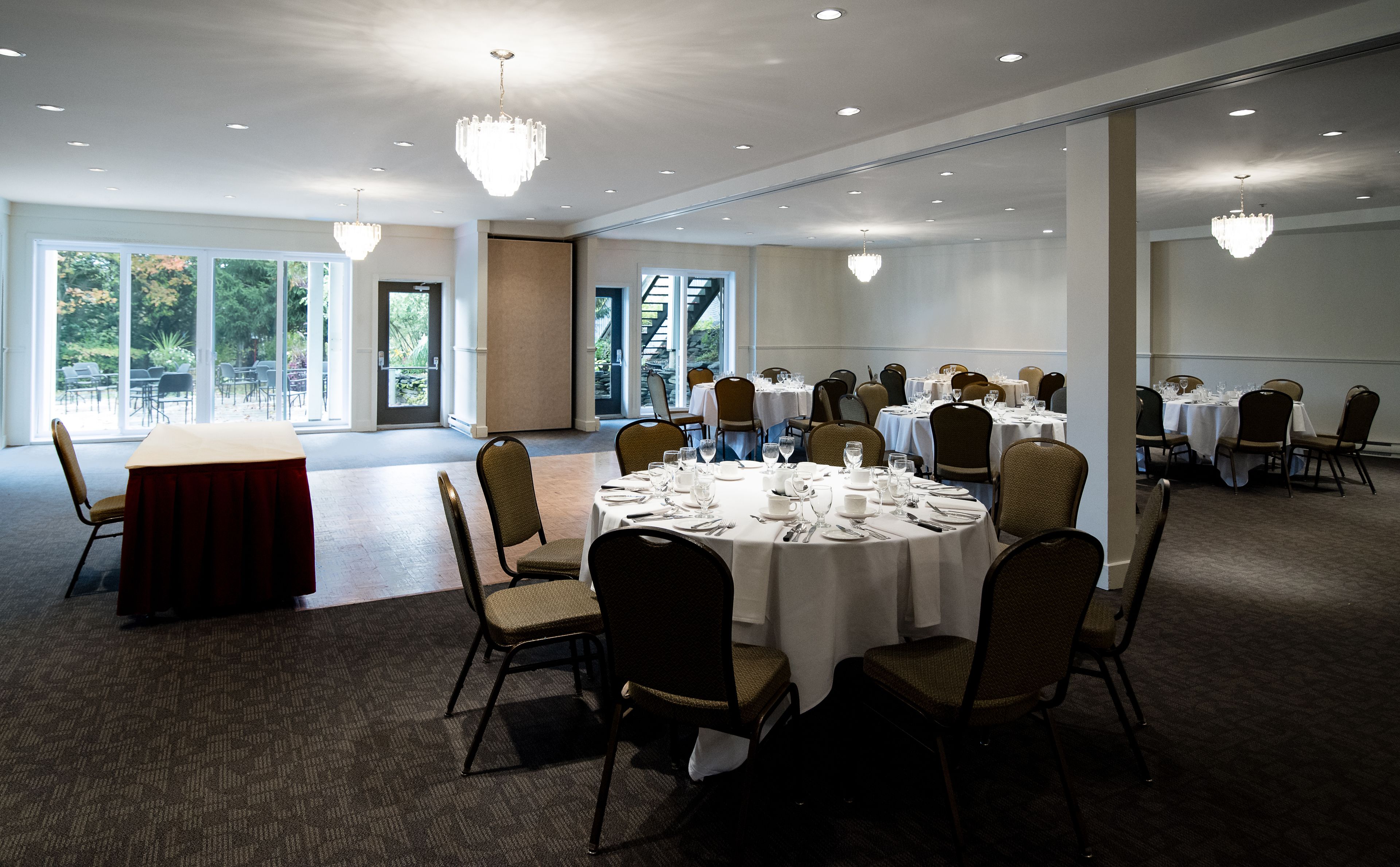 A banquet hall with round tables set for dining, surrounded by chairs, and large windows providing natural light.
