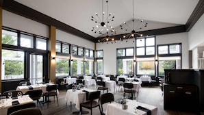 A spacious dining area with multiple tables set for dining, large windows offering views of the outdoors, and modern lighting fixtures.