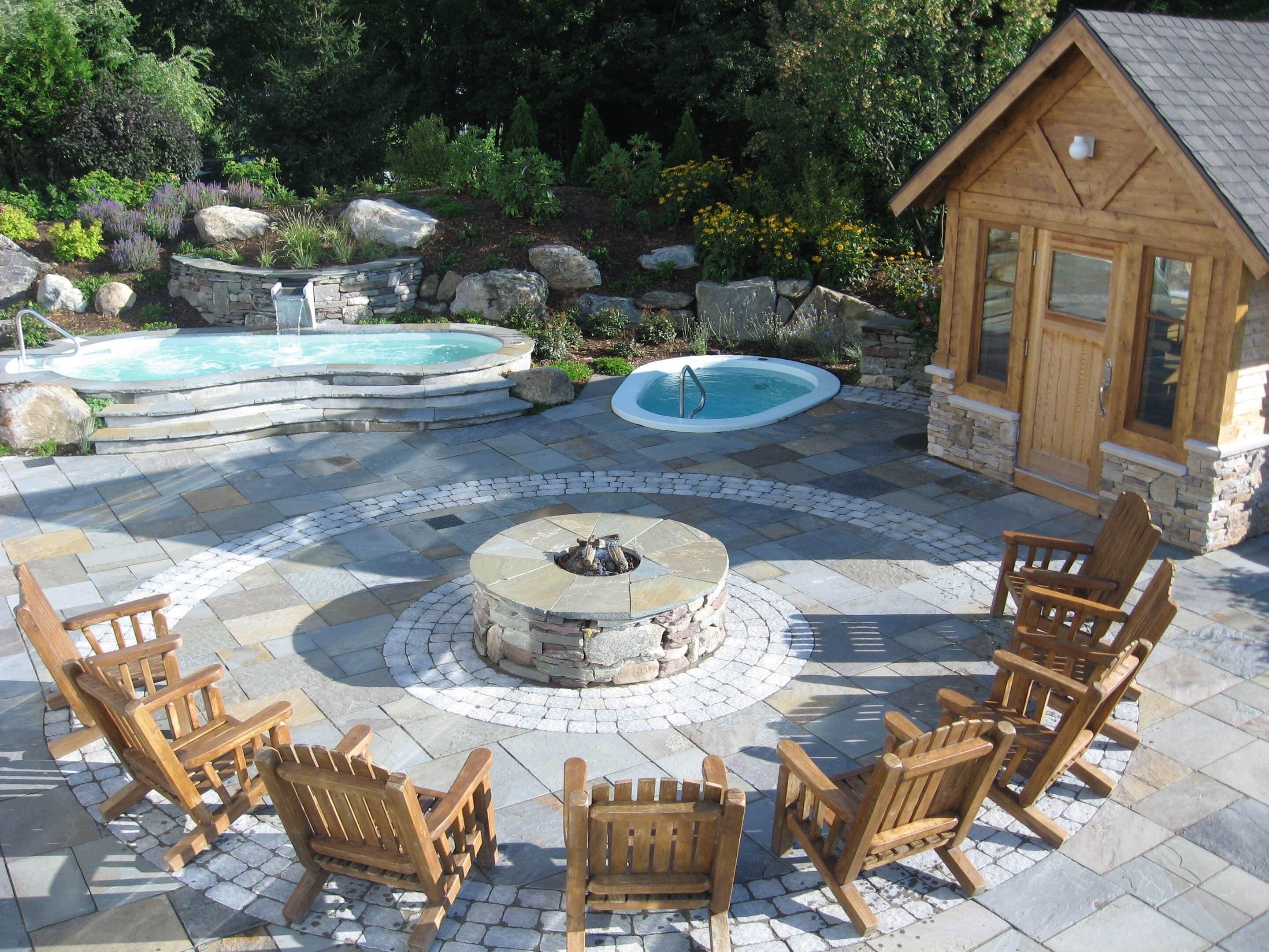 A patio with a stone fire pit, wooden chairs, and two hot tubs.