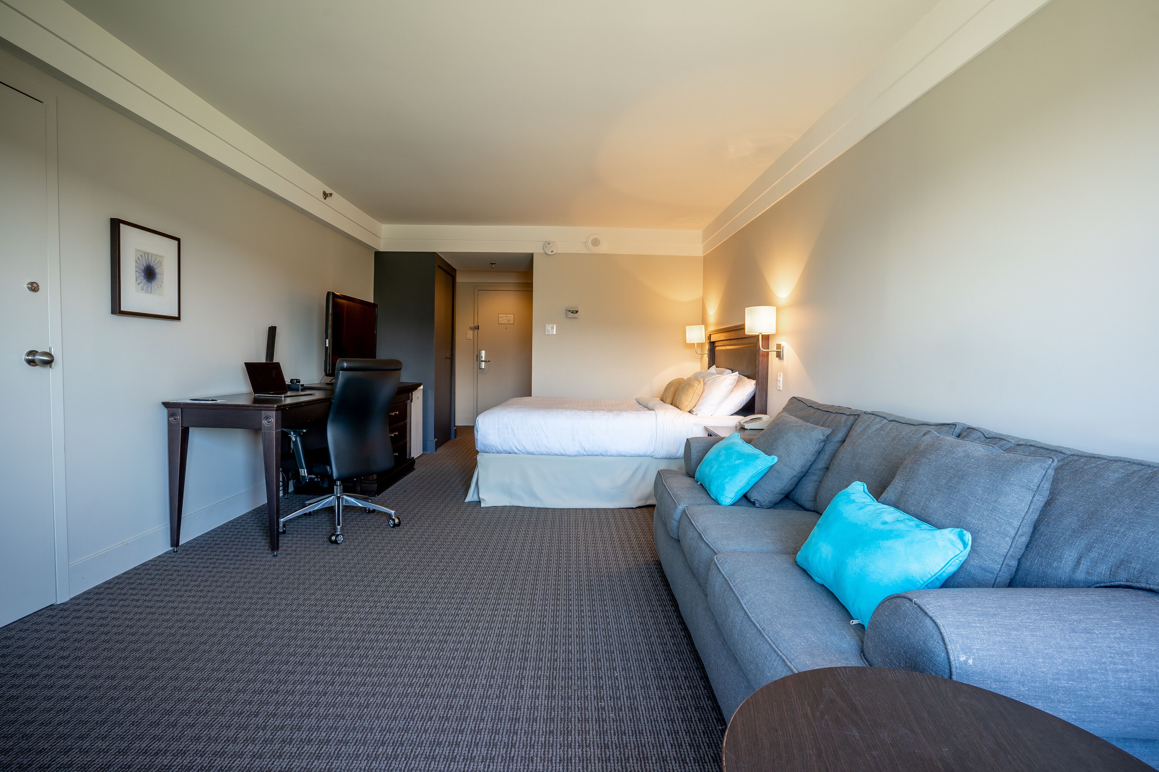 A hotel room with a grey carpet, a grey sofa with blue cushions, a white bed, a desk with a chair, and a framed picture on the wall.