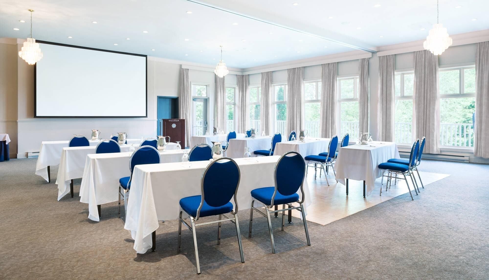 A conference room with tables, chairs, a projector screen, and large windows.