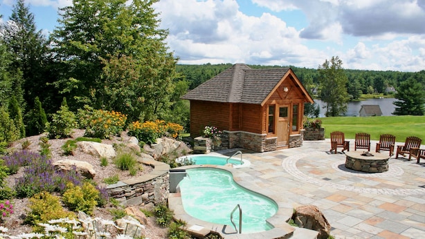 A wooden cabin with a swimming pool and outdoor seating area surrounded by a landscaped garden and a lake view.