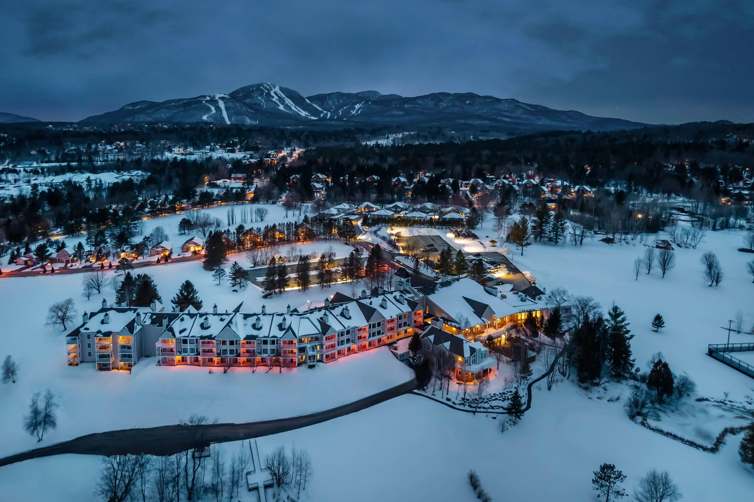 A snow-covered resort with multiple buildings, a ski slope, and a mountain in the background.