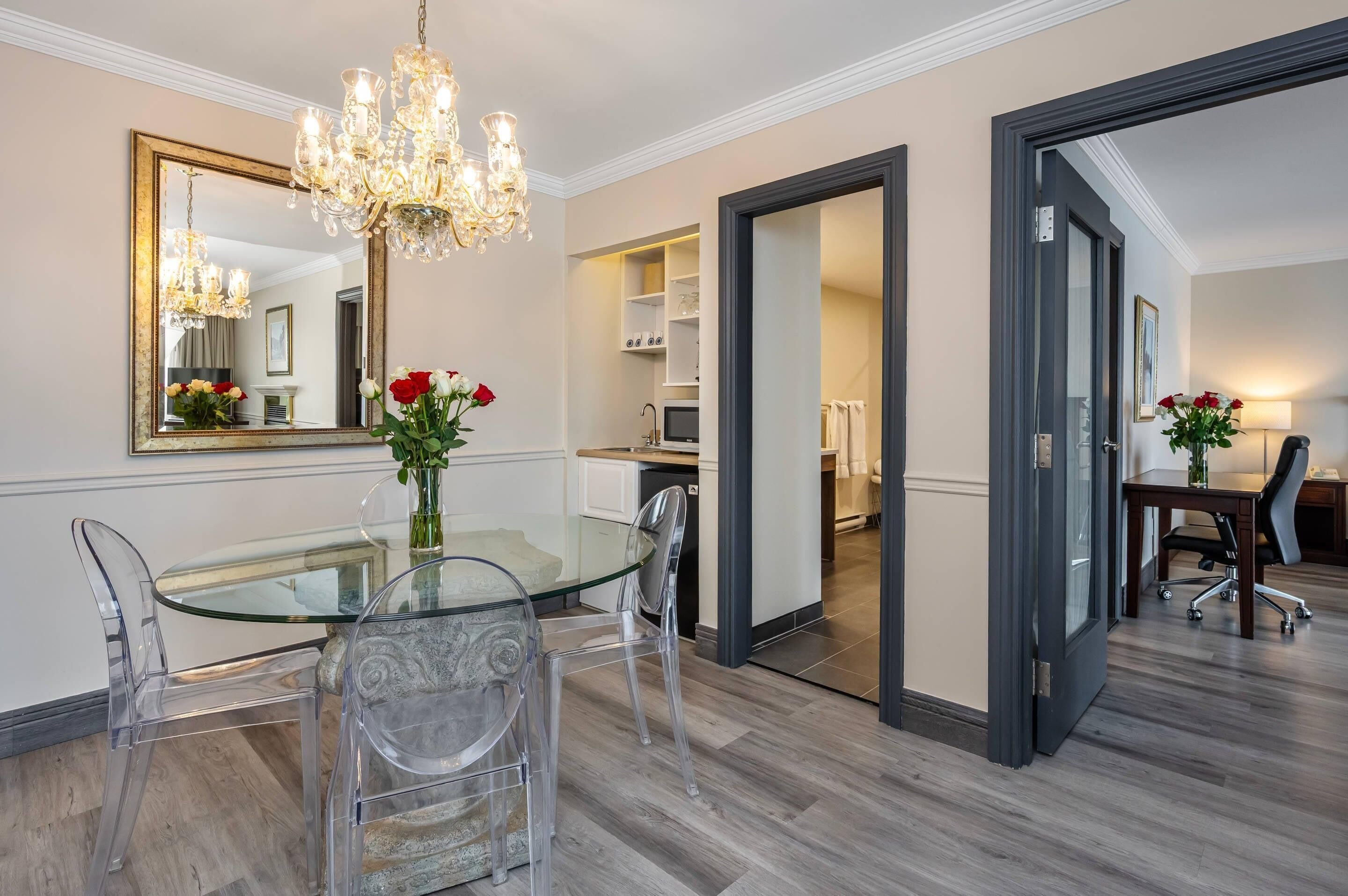 A dining area with a glass table, clear chairs, a chandelier, and a mirror.