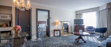 A hotel room with a chandelier, a dining table, a sofa, a TV, and a fireplace.