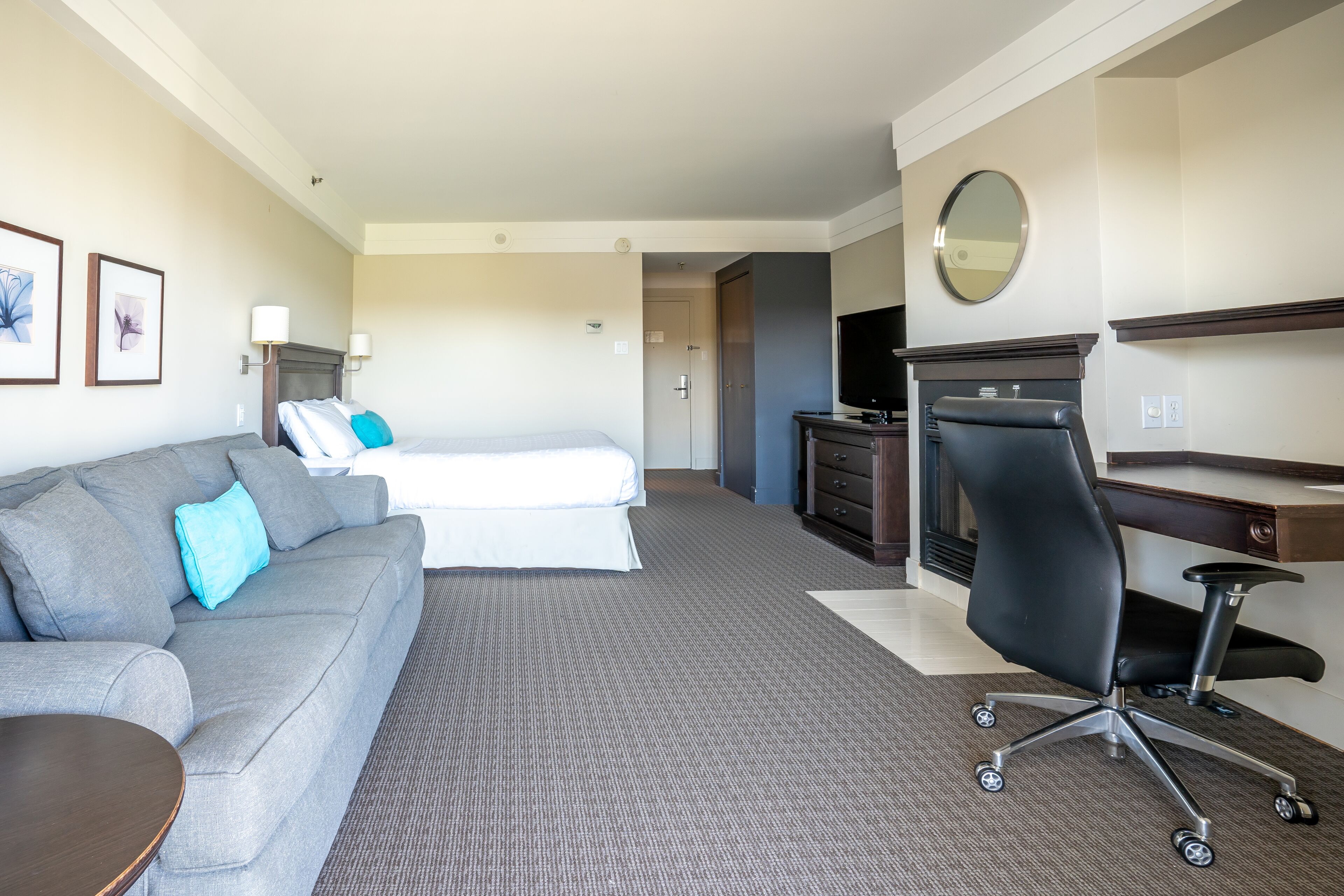 A hotel room with a grey sofa, a bed with white bedding, a desk with a chair, a television, and framed pictures on the wall.
