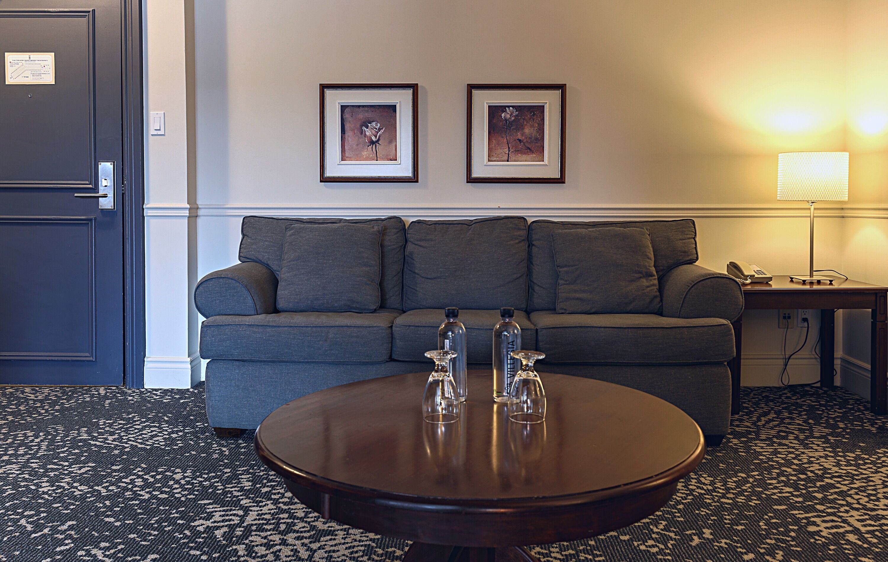 A hotel room with a grey sofa, two framed pictures on the wall, a round wooden coffee table with three bottles, and a lamp on a side table.