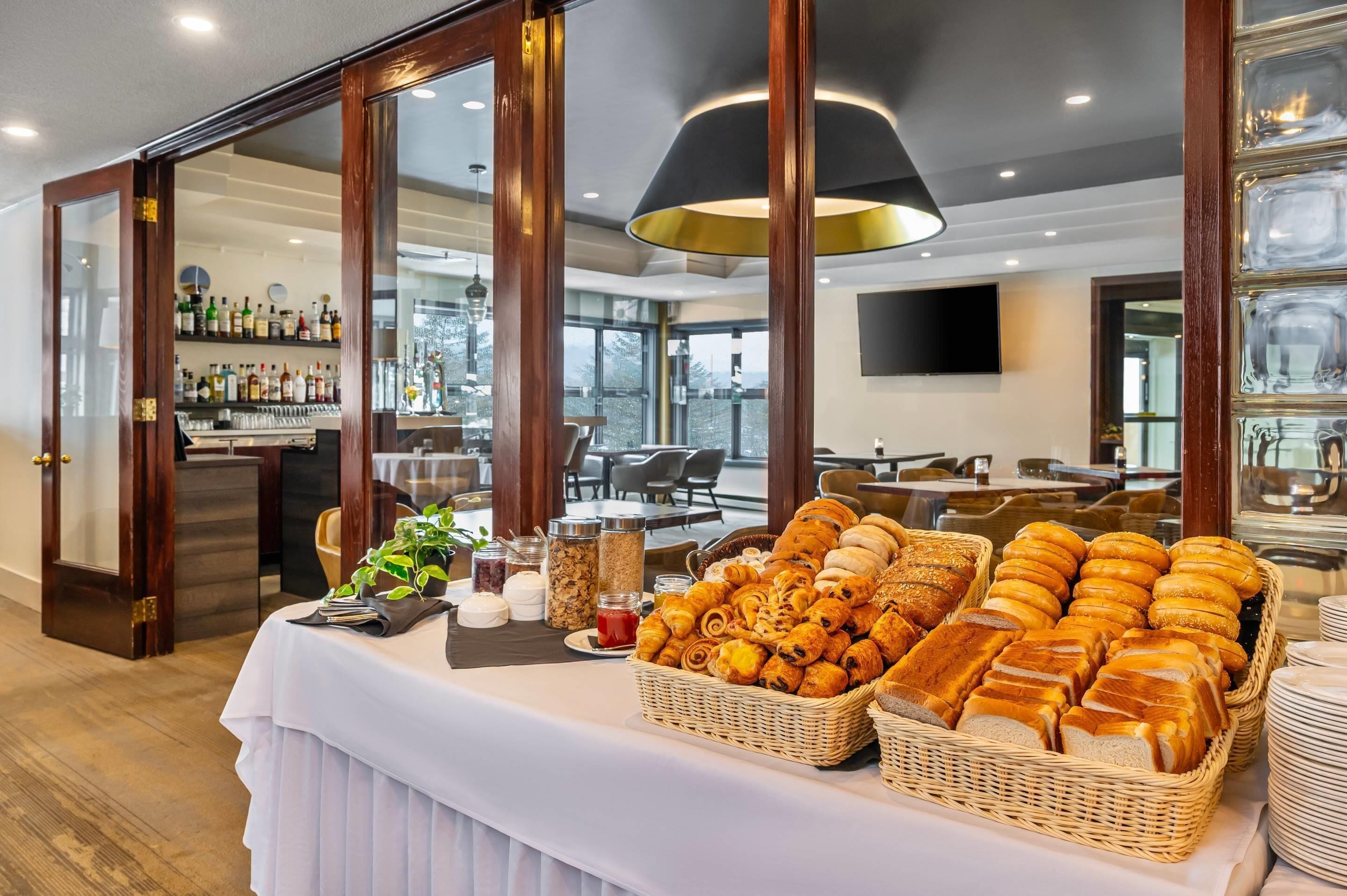 A breakfast buffet with various breads and pastries, a bar area with liquor bottles, and a view of the outdoors through large windows.