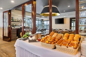 Un bufé de desayuno con panes y pasteles variados, una zona de bar con botellas de licores y vistas al exterior a través de amplias ventanas.