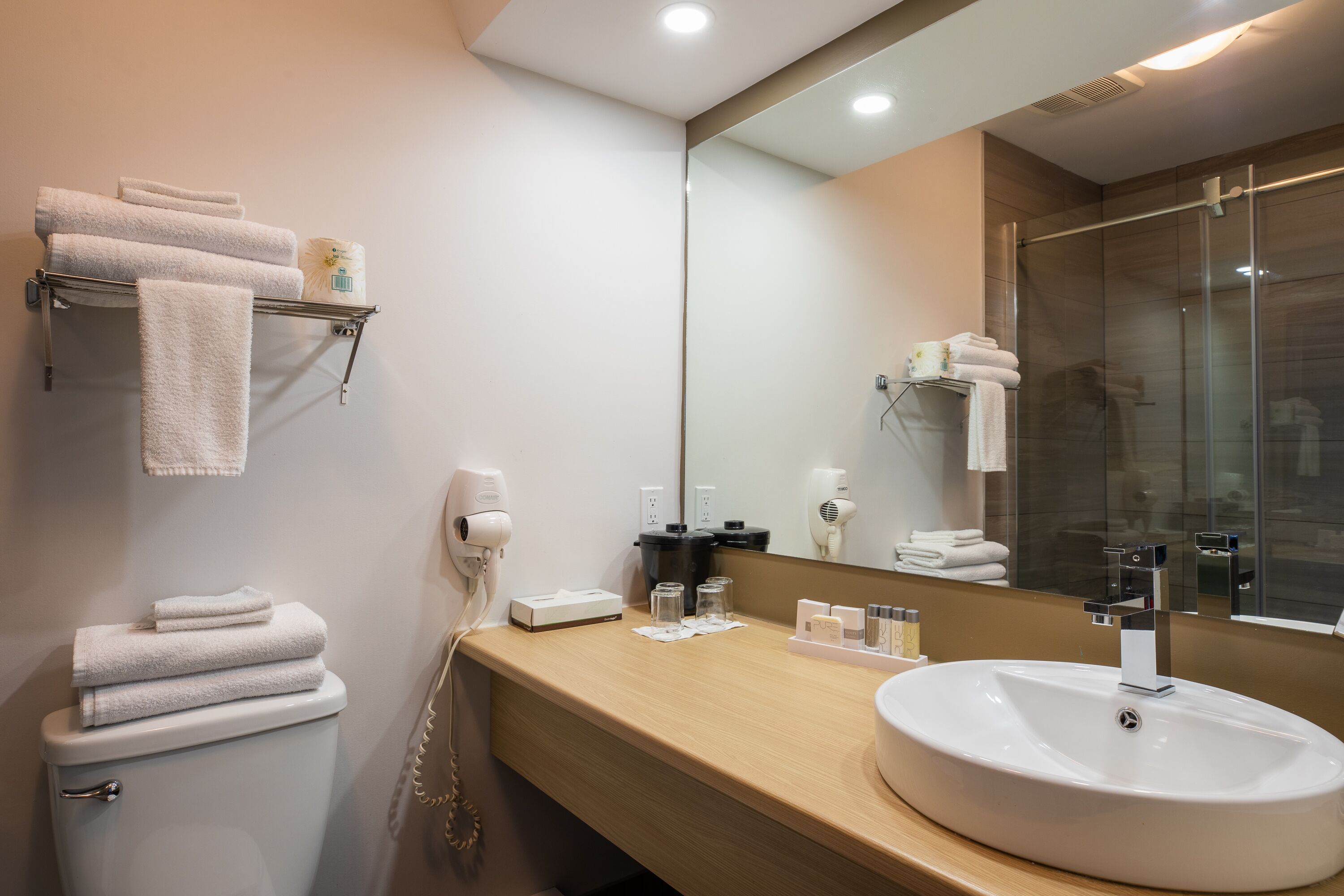 A modern bathroom with a toilet, sink, and large mirror. There are towels on a shelf and a hairdryer on the wall.