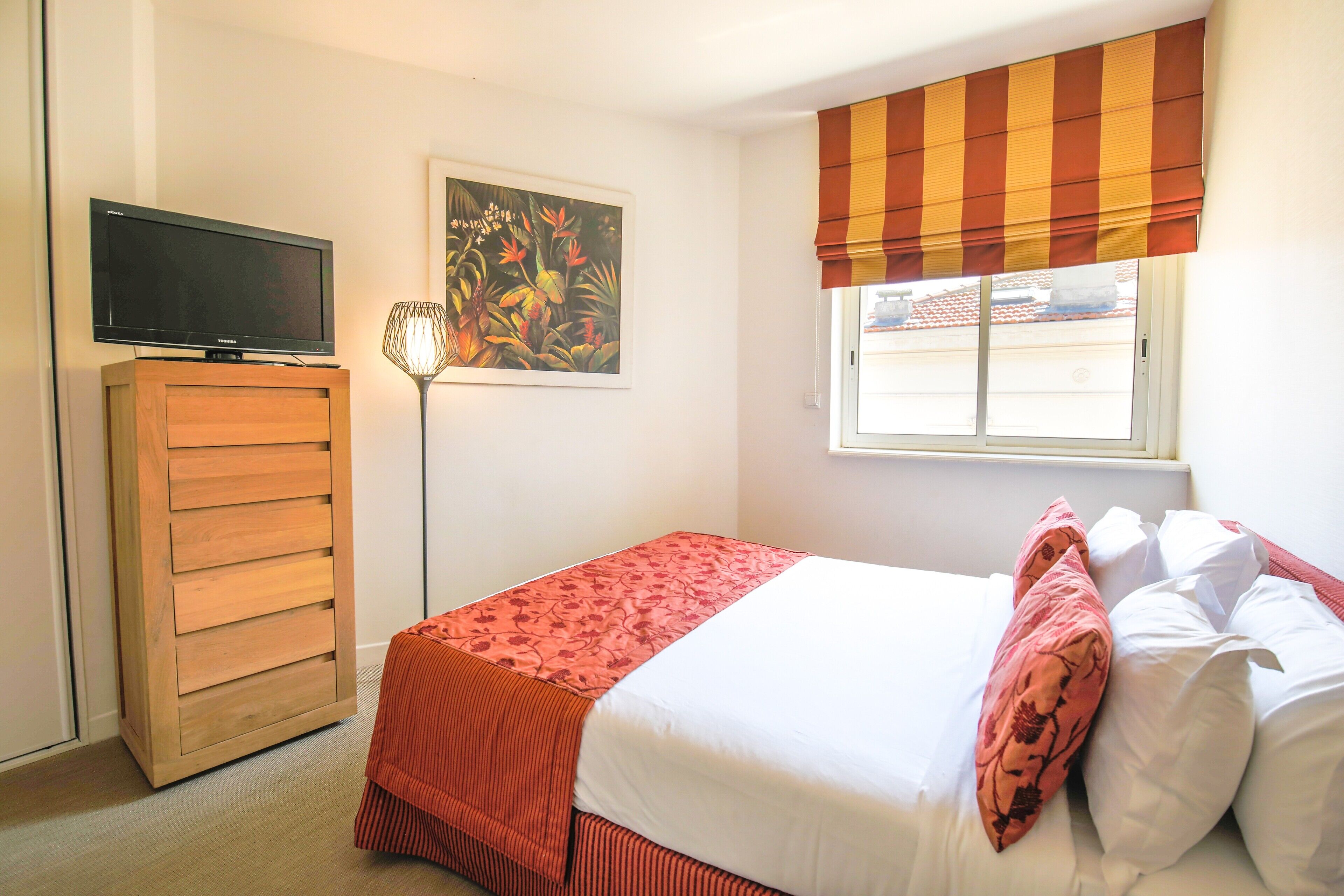 A bedroom with a bed, a TV on a wooden cabinet, a painting on the wall, and a window with a striped blind.