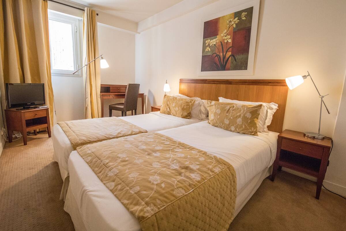 Habitación de hotel con dos camas, un escritorio con televisor, una silla y dos lámparas de noche.