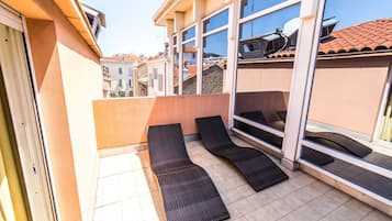 Un balcón con dos sillones reclinables de color oscuro, suelo de baldosas y ventanas amplias que dan vistas a edificios y una colina.