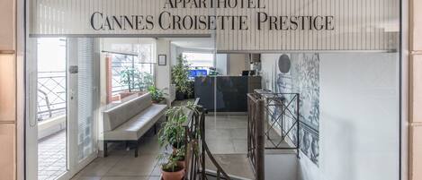 Interior do Appartôtel Cannes Croisette Prestige, com área de recepção, um banco, plantas em vasos e uma grande obra de arte emoldurada na parede.