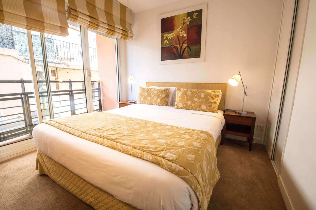 Habitación de hotel con una cama grande, una mesita de noche, una lámpara, un cuadro en la pared y vistas por la ventana.