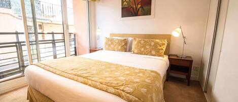 A hotel room with a large bed, a nightstand, a lamp, a painting on the wall, and a view through the window.