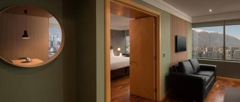 A modern hotel room with a wooden floor, a round mirror, a black leather sofa, a bed, and a TV.