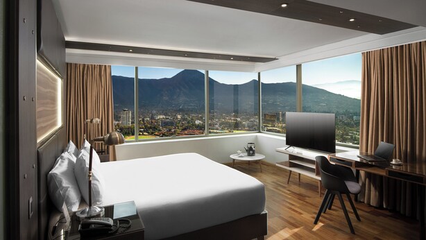 Quarto de hotel moderno com uma cama grande, uma escrivaninha, televisão e vista para a cidade e montanhas.
