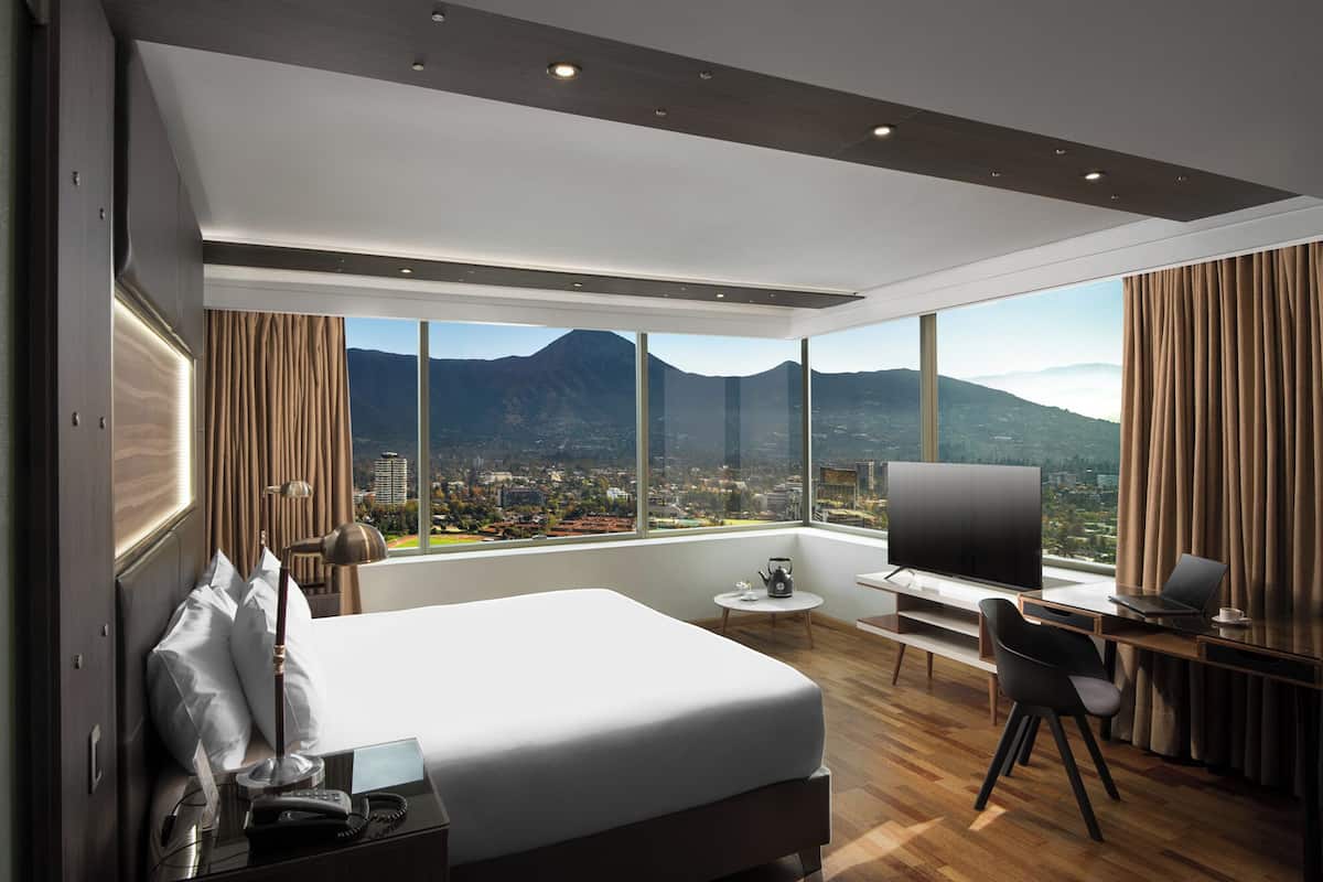 A modern hotel room with a large bed, a desk, a TV, and a view of a city and mountains.