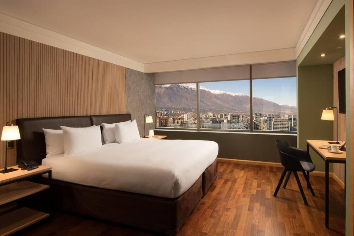 A hotel room with a large bed, a desk, and a view of a cityscape and mountains.