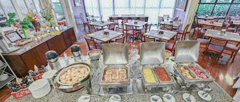 Montaje de bufé con distintas estaciones de comida, entre ellas una barra de ensaladas y una de carnes, en un amplio comedor con numerosas mesas y sillas.