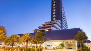 Un hotel moderno con una torre central alta, varios pisos y un estacionamiento con coches y palmeras.