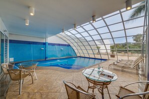 An indoor swimming pool with a glass ceiling, surrounded by wicker chairs and a glass table. - Jk Premium Hotel e Eventos (Londrina)
