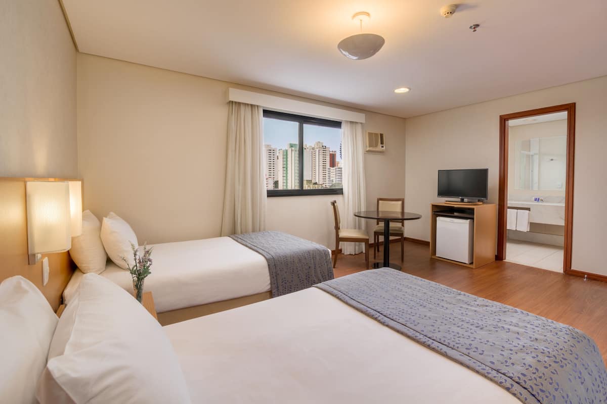 Habitación de hotel con dos camas, un escritorio, una silla, un televisor y un ventanal con vista a la ciudad.