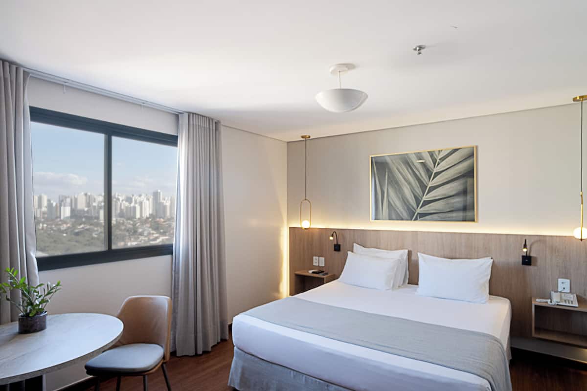 Una habitación de hotel moderna con una cama grande, un escritorio con silla, una planta en maceta y vistas a la ciudad desde la ventana.