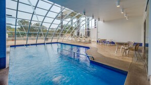 Una piscina interior con paredes de vidrio transparente, sillones de descanso y suelo de baldosas.