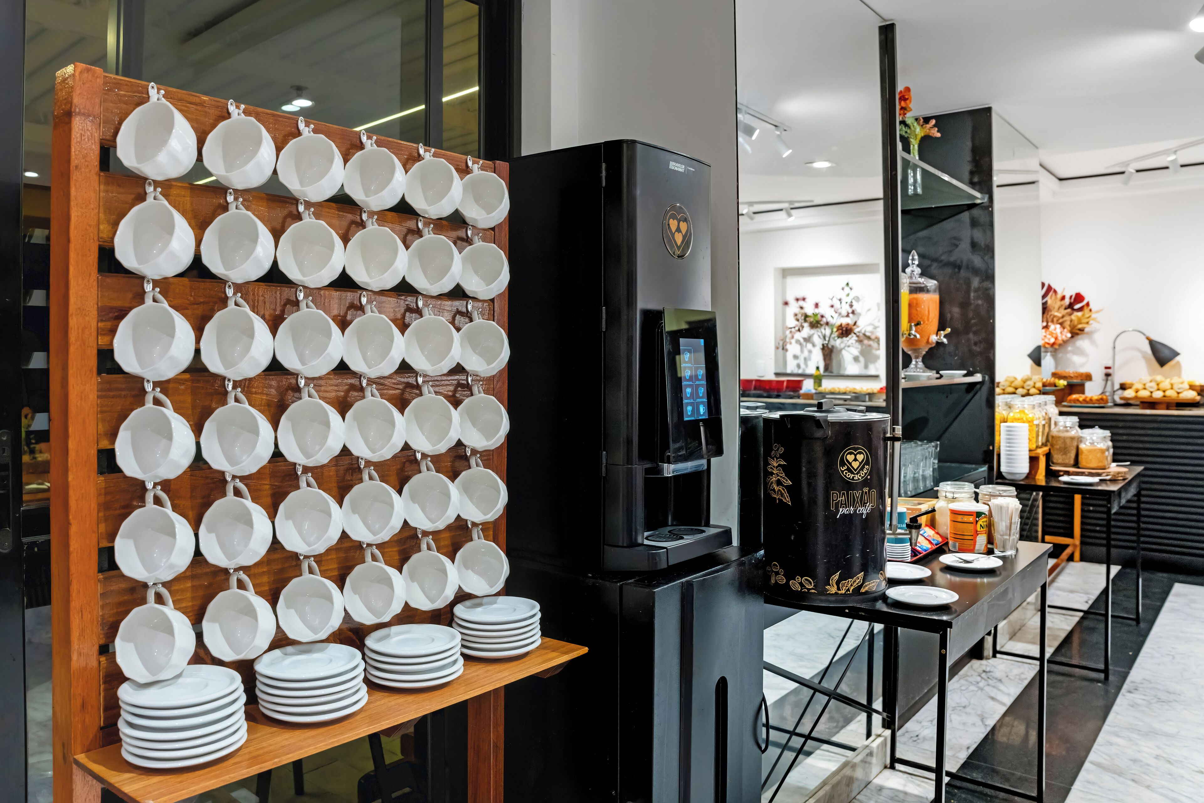 A coffee station with a coffee machine, a display of white cups and plates, and a counter with various food items.