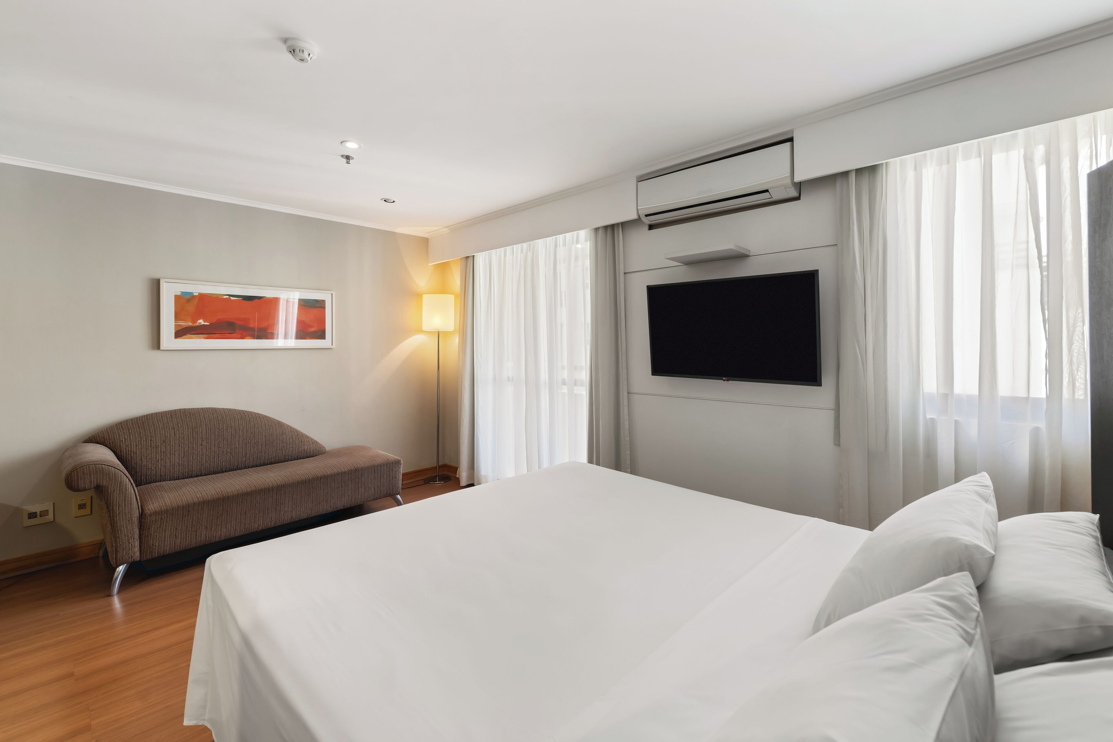 A hotel room with a large bed, a sofa, a flat-screen TV, and a window with curtains.