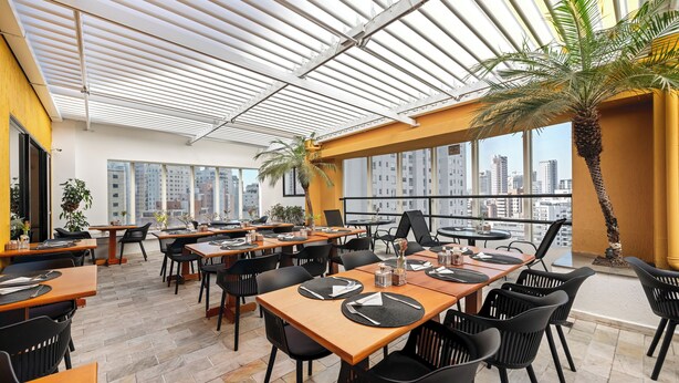 A modern restaurant with high tables and black chairs, large windows, and a city view. - SLAVIERO São Paulo Ibirapuera (São Paulo)