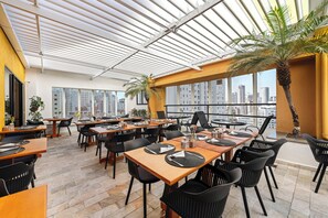 Um restaurante moderno com mesas altas e cadeiras pretas, amplas janelas e vista para a cidade.