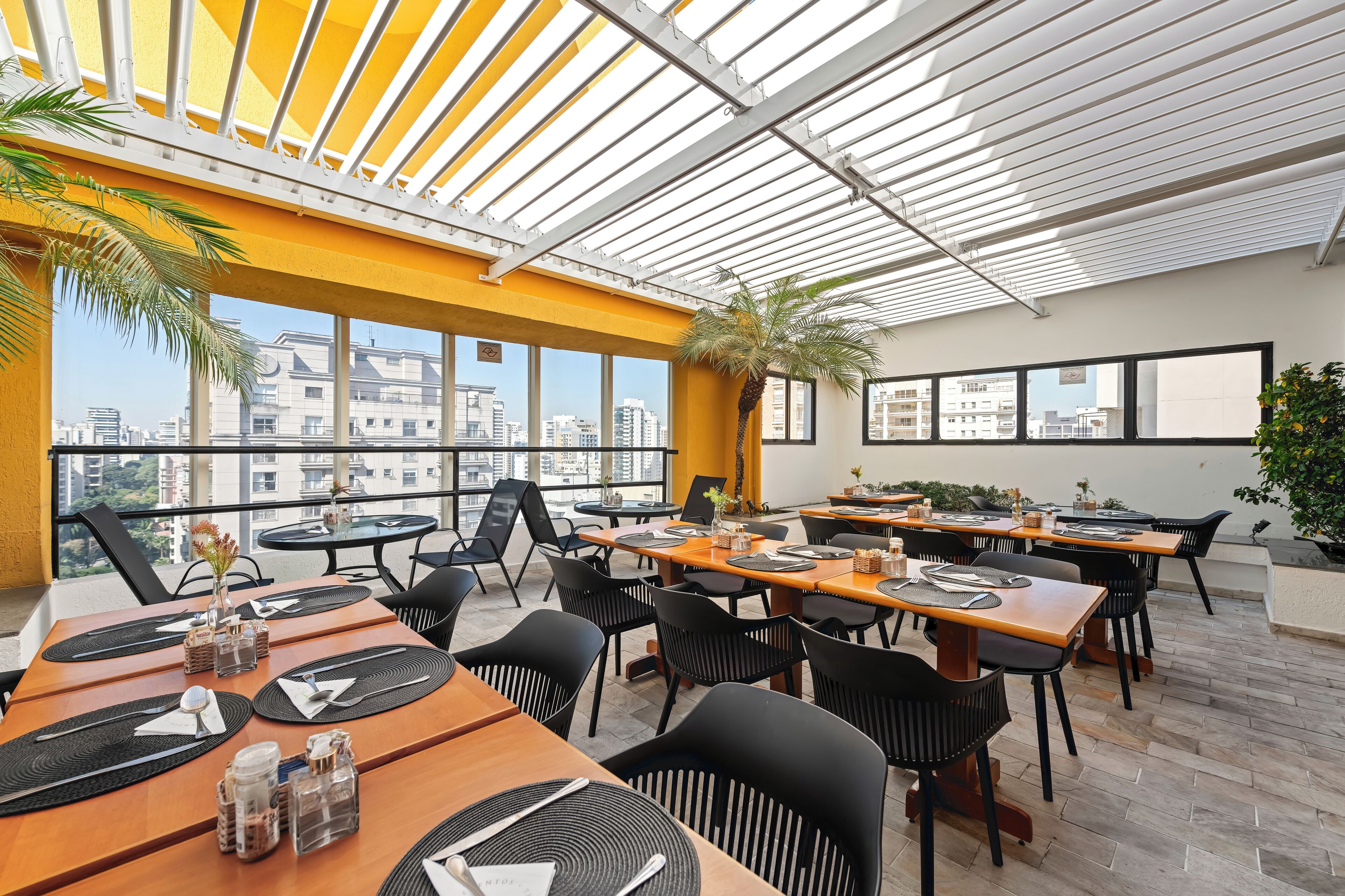 Un espacio de comedor en la azotea con varias mesas y sillas, amplias ventanas que ofrecen vistas de la ciudad y un diseño arquitectónico moderno.
