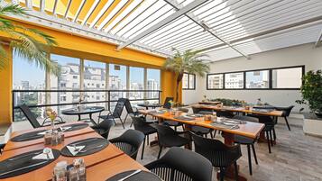 Un espacio de comedor en la azotea con varias mesas y sillas, amplias ventanas que ofrecen vistas de la ciudad y un diseño arquitectónico moderno.