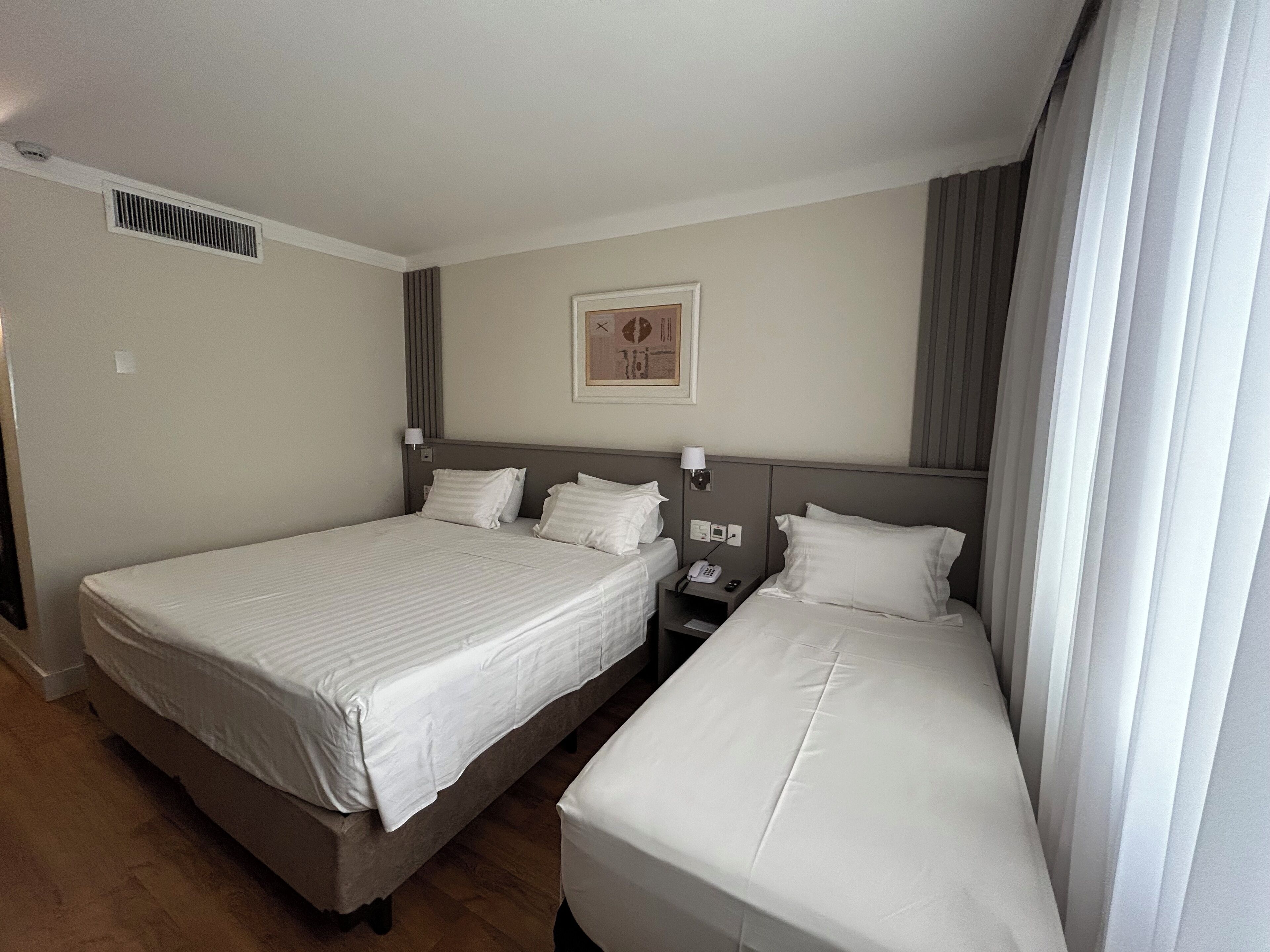 A hotel room with two beds, a nightstand with a phone, a wall-mounted light, and a vent.