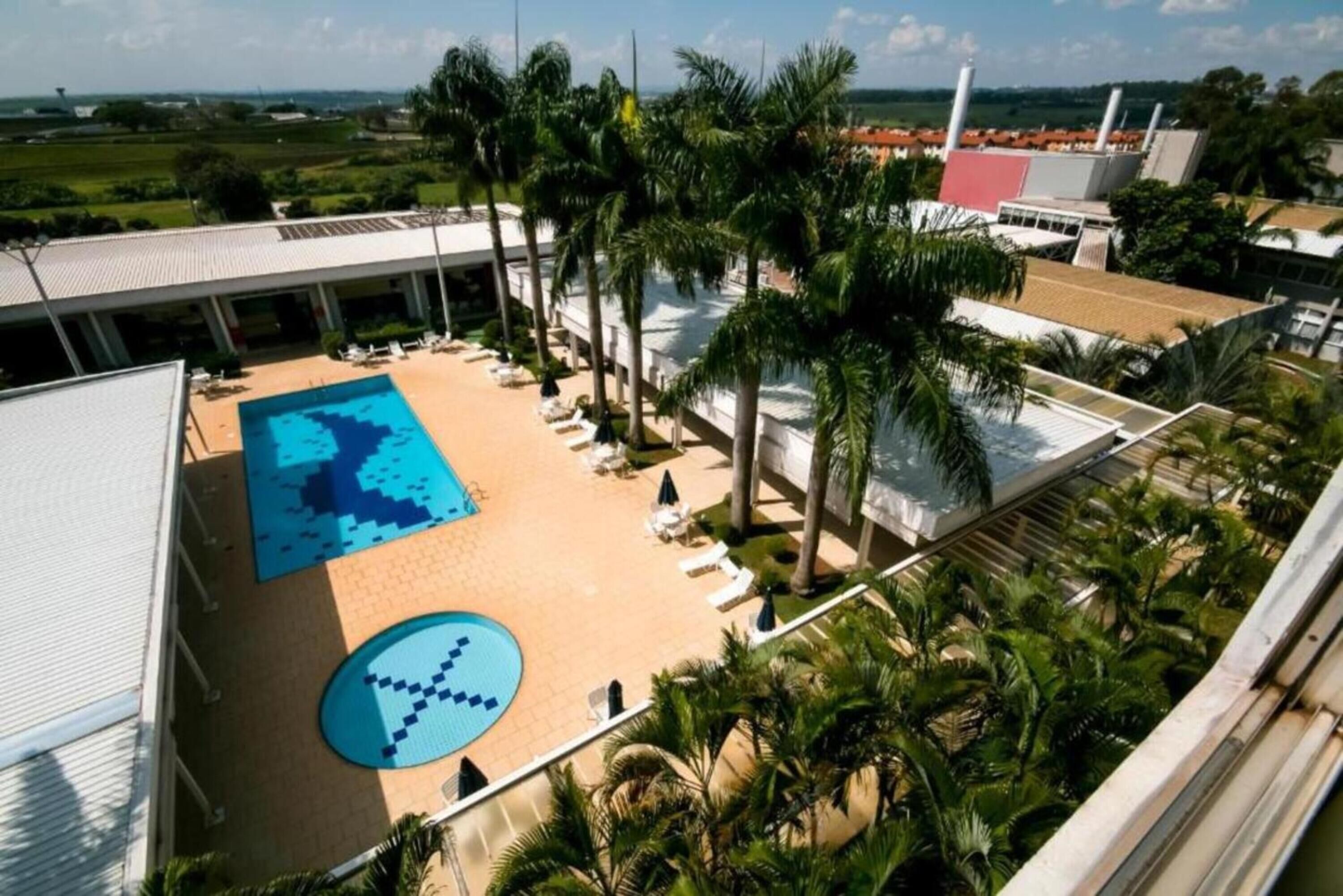 A rooftop pool area with a rectangular pool and a circular pool, surrounded by palm trees and lounge chairs.