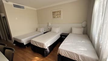 A hotel room with three single beds, a small table, a chair, and a painting on the wall.