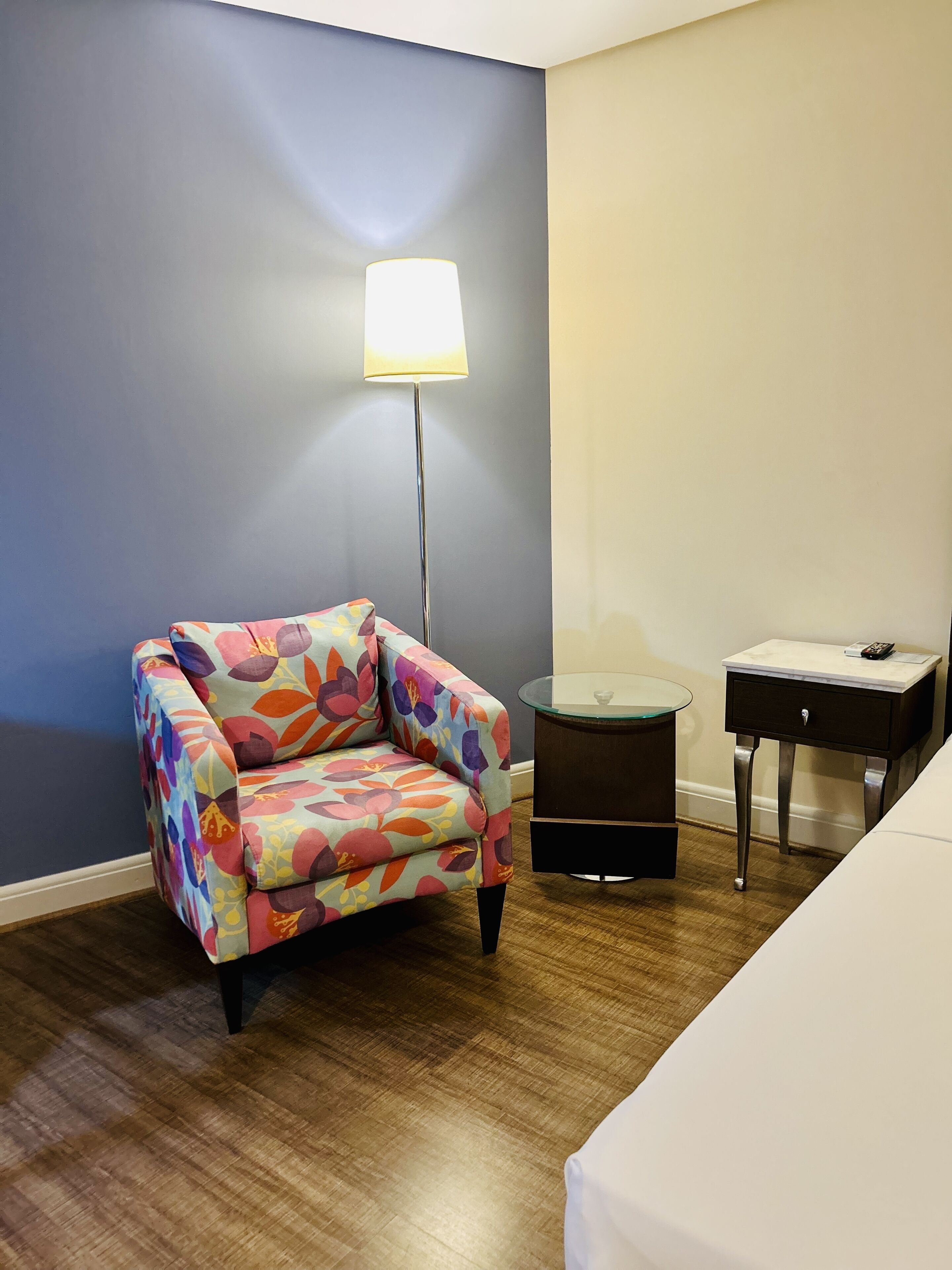 Ein Zimmer mit einem bunten Sessel, einem Nachttisch, einer Lampe und einem Bett.