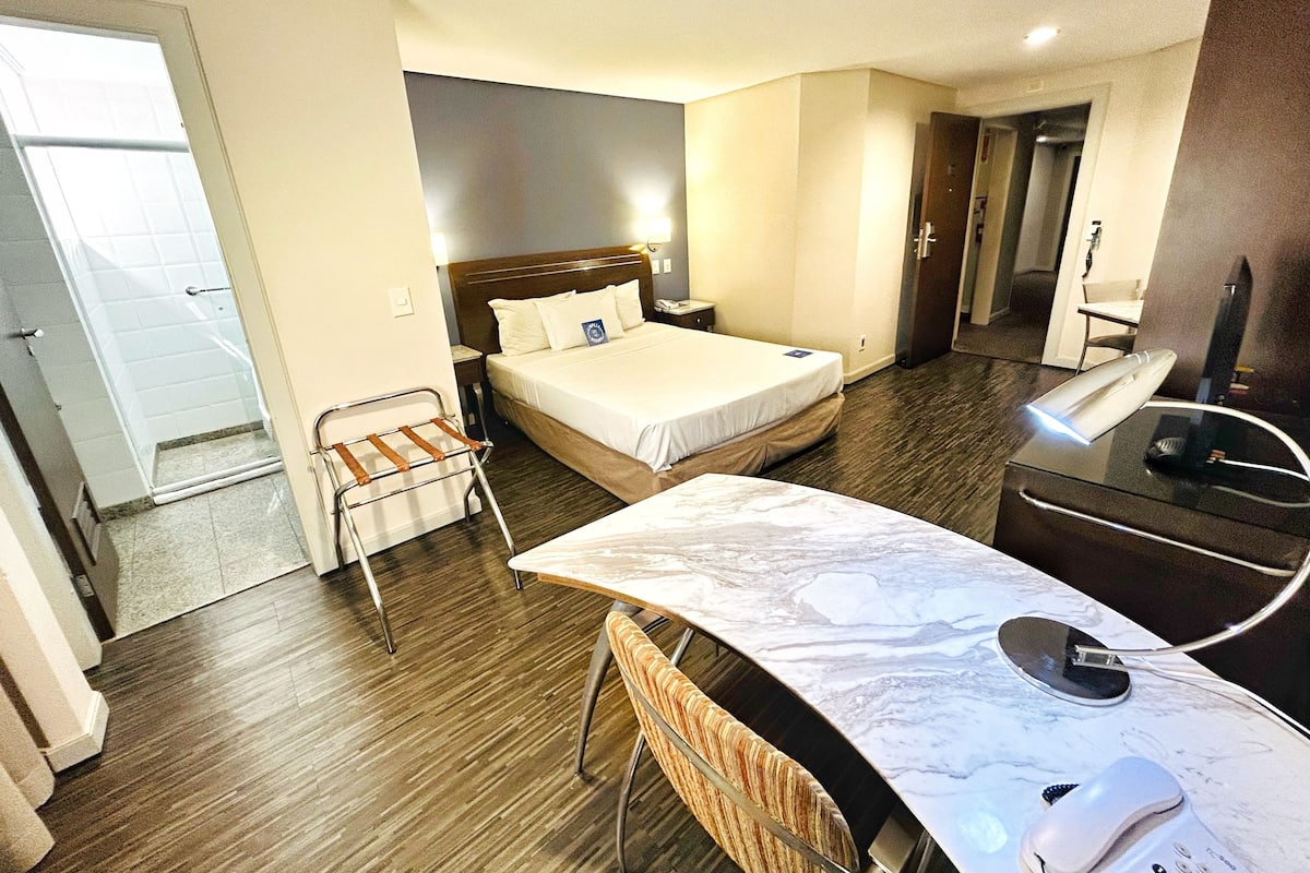 A hotel room with a bed, bedside table, desk with a computer, and a chair.