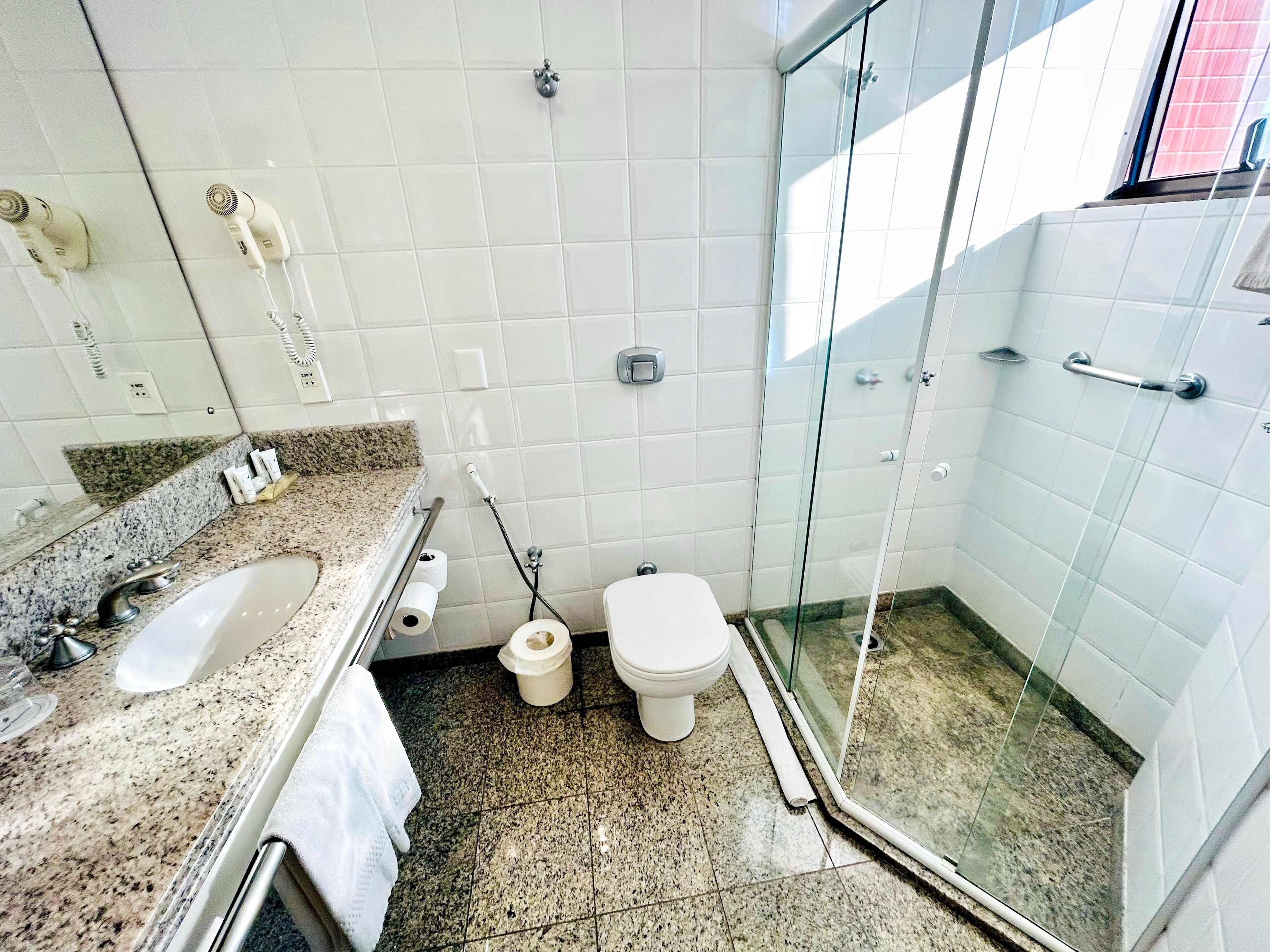 Un baño con encimera de granito, lavamanos blanco, inodoro y zona de ducha con mampara de vidrio.