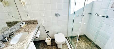 Un baño con encimera de granito, lavamanos blanco, inodoro y zona de ducha con mampara de vidrio.